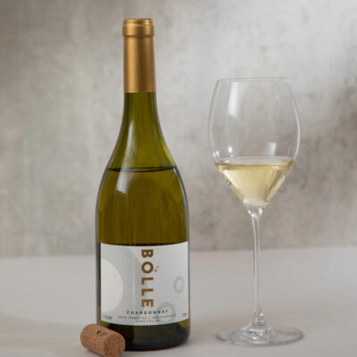 Flaska af BOLLE Still Chardonnay, korktappi og glas með hvítvíni við ljósan steinvegg